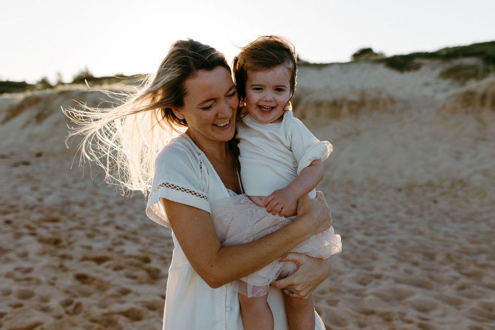 A joyful mother in a white dress holds her laughing toddler on a sunny sandy beach.