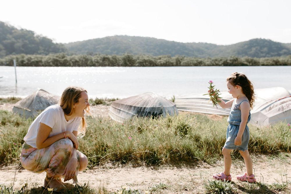A mother crouches as her toddler daughter offers her a small plant near a scenic lakeside with mountains behind.