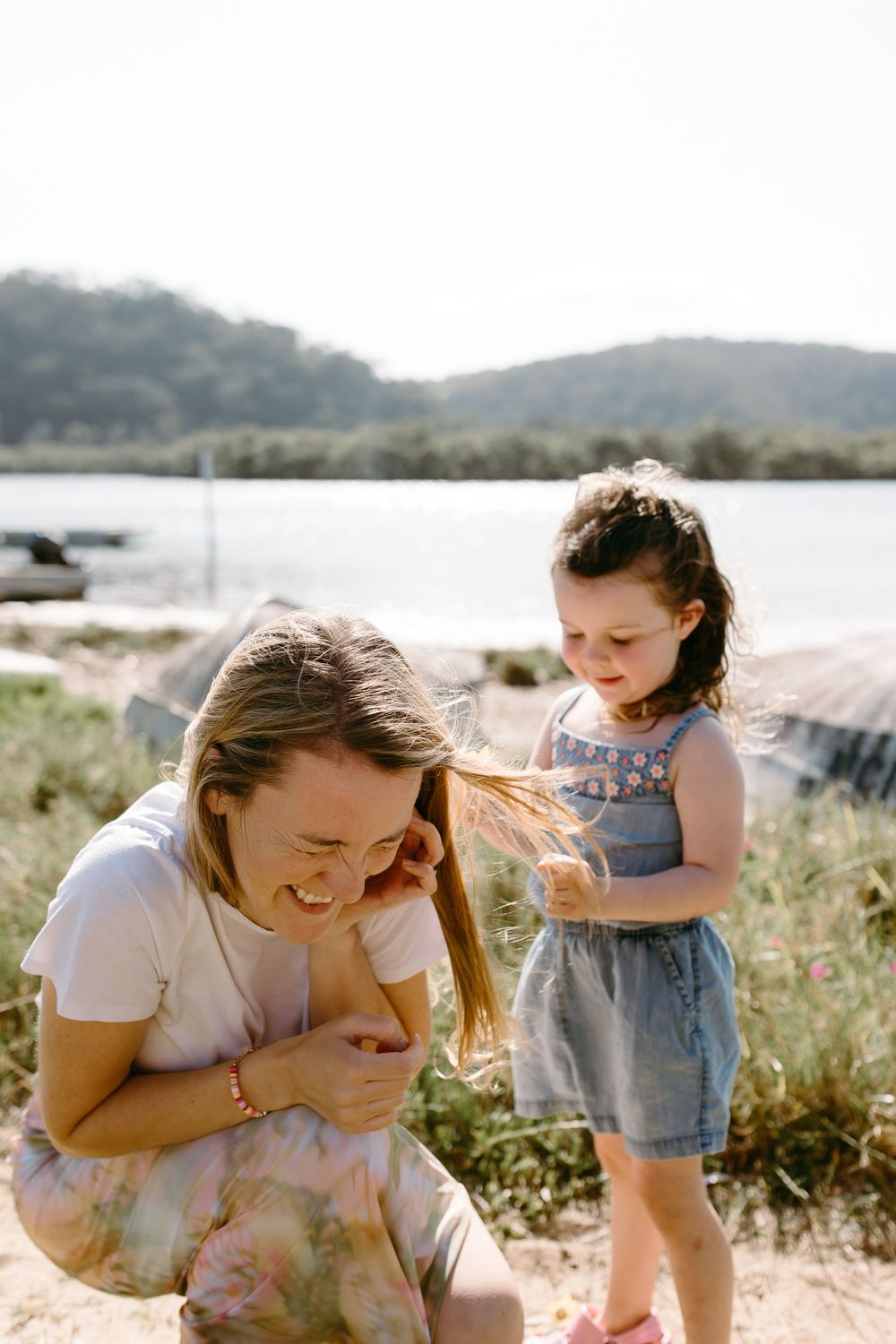 A woman crouches laughing as a young girl in a denim dress plays with her hair near a sunny lakeside.