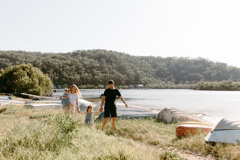 Family walking along a scenic lakeside shore with lush green hills and boats in the background on a sunny day.