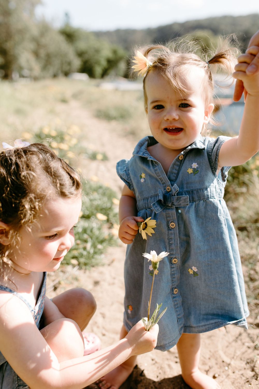 Two young girls in denim dresses play outdoors, holding white daisies in a sunny field.