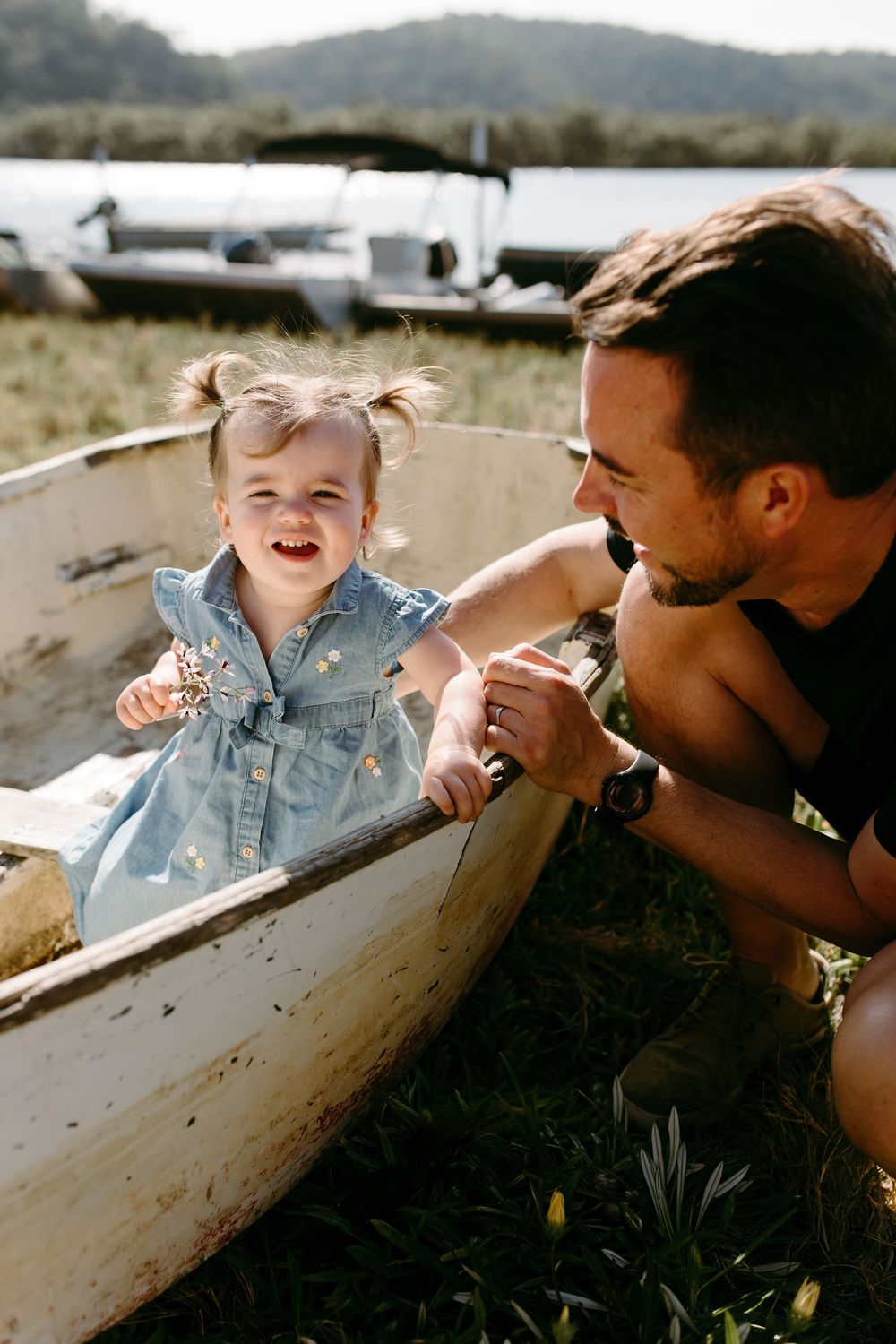 A joyful toddler girl in a denim dress sits in an old white boat while her father smiles at her outdoors.