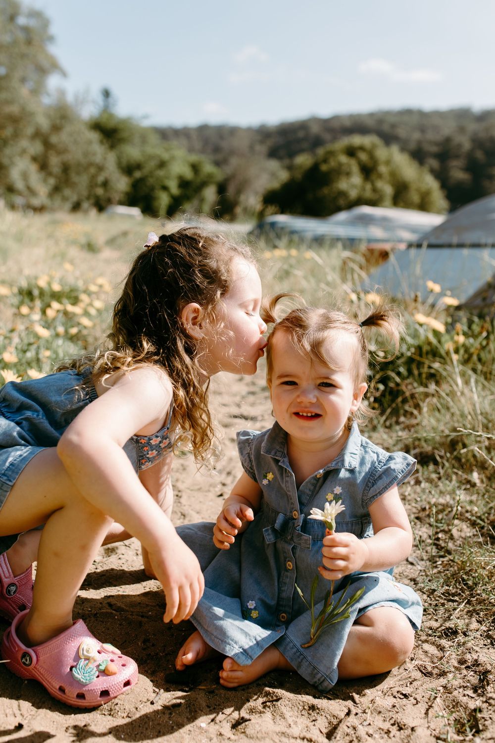 Two young sisters in denim outfits sit outdoors, older girl kissing younger sibling holding wildflowers in sunny field.