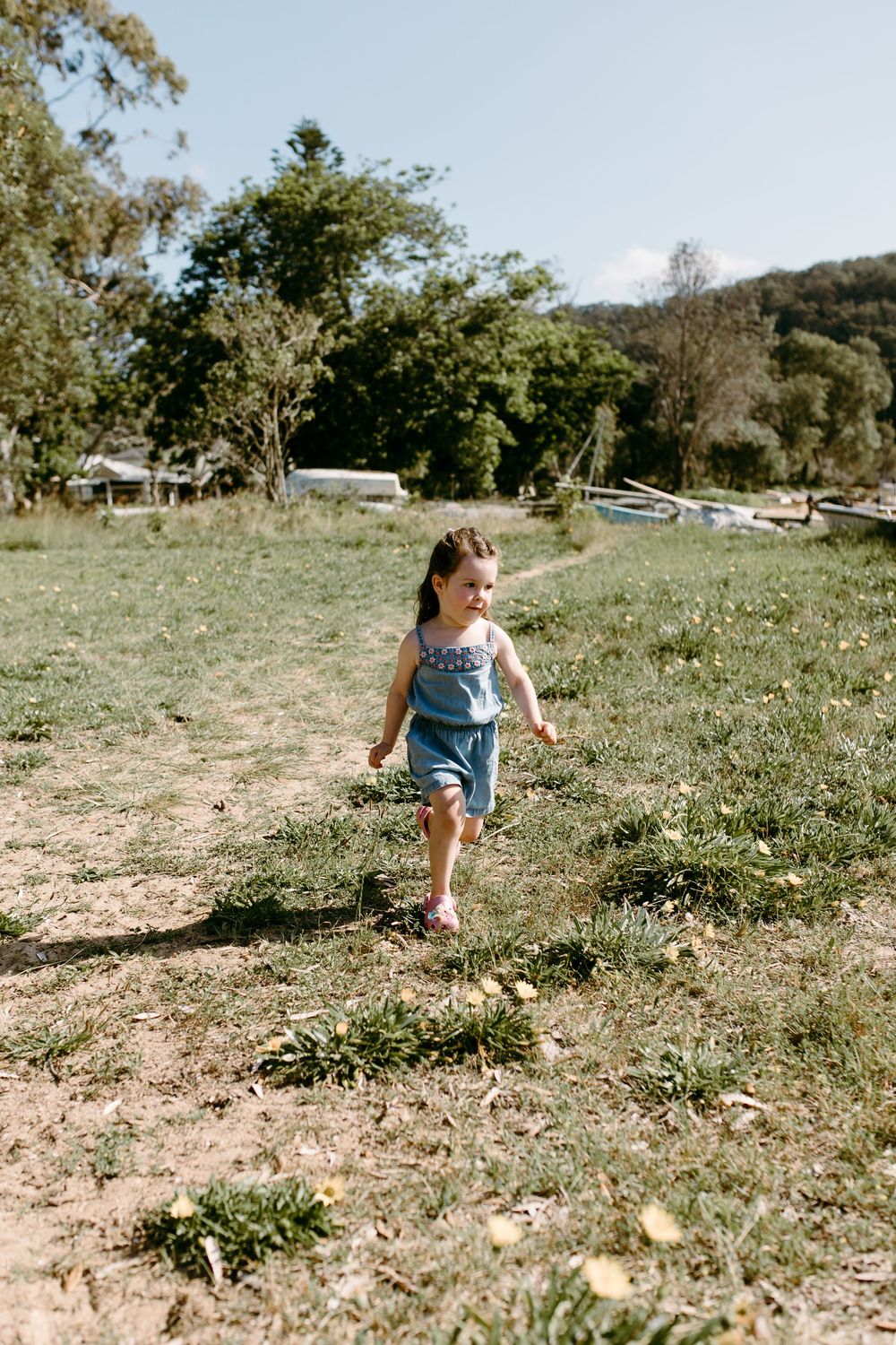 A young girl in denim overalls runs barefoot across a sunny green field with trees in the background.