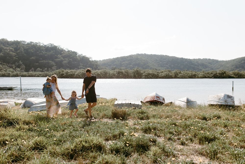 Family of four walking hand-in-hand along a scenic lakeside shore with green hills and moored boats in background.