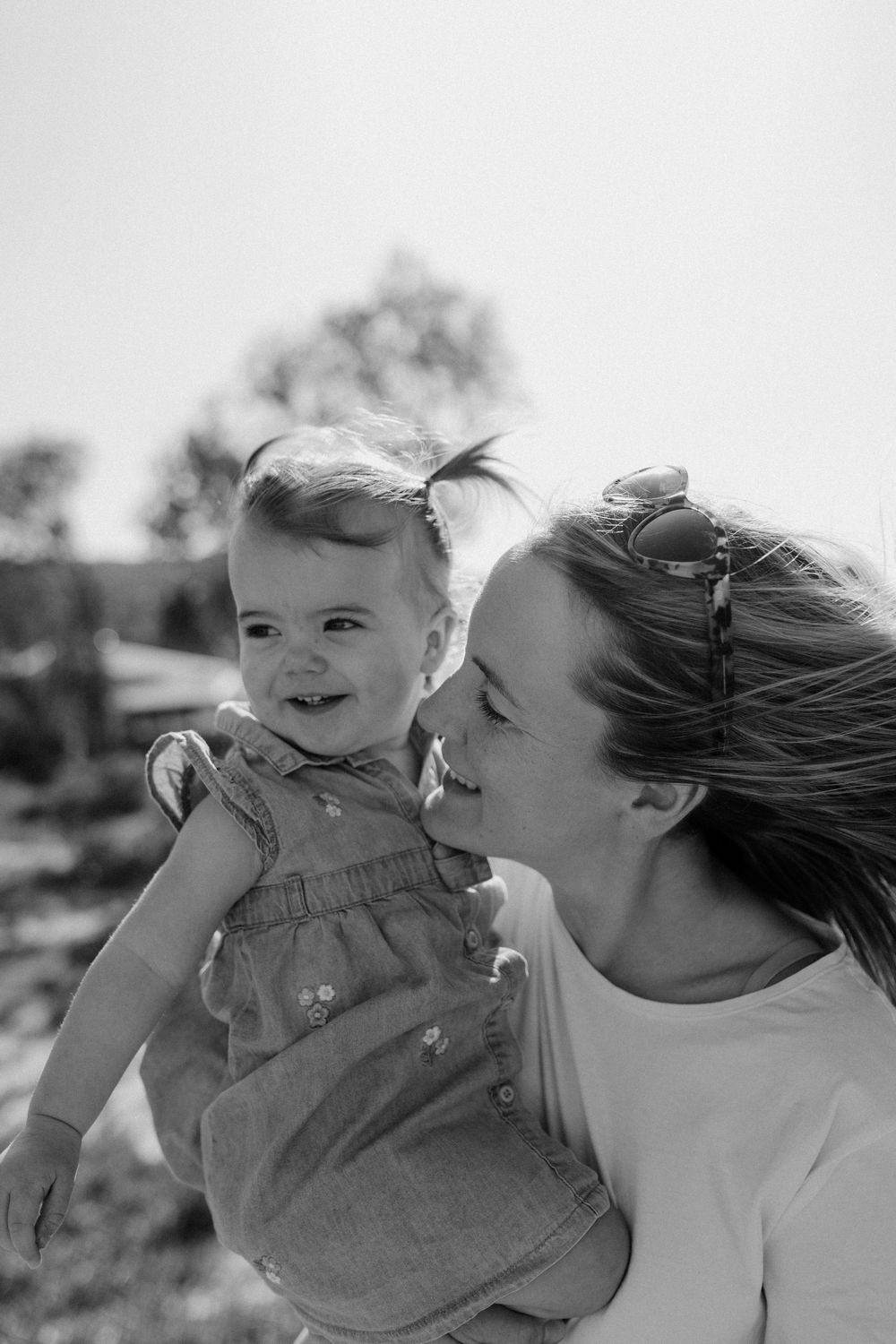 Black and white photo of a smiling baby in a dress being held by a loving mother outdoors.
