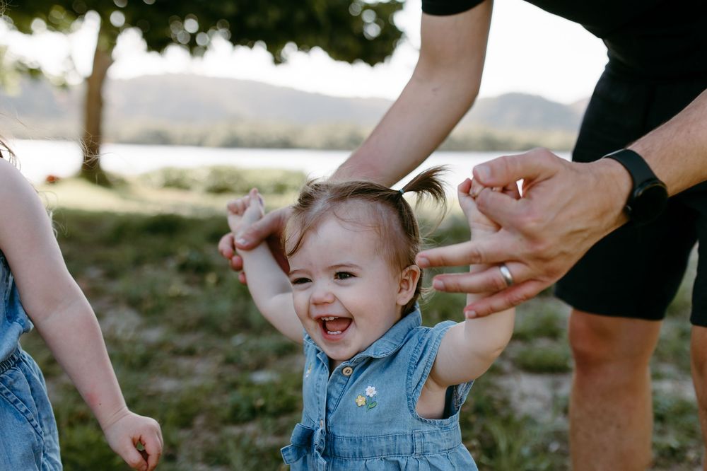 Laughing baby girl in denim overalls being held up by parents outdoors near a lake.