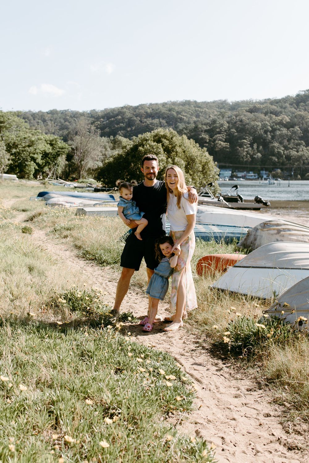 Family of four posing on a sandy path by the waterfront with boats and lush green hills in the background.