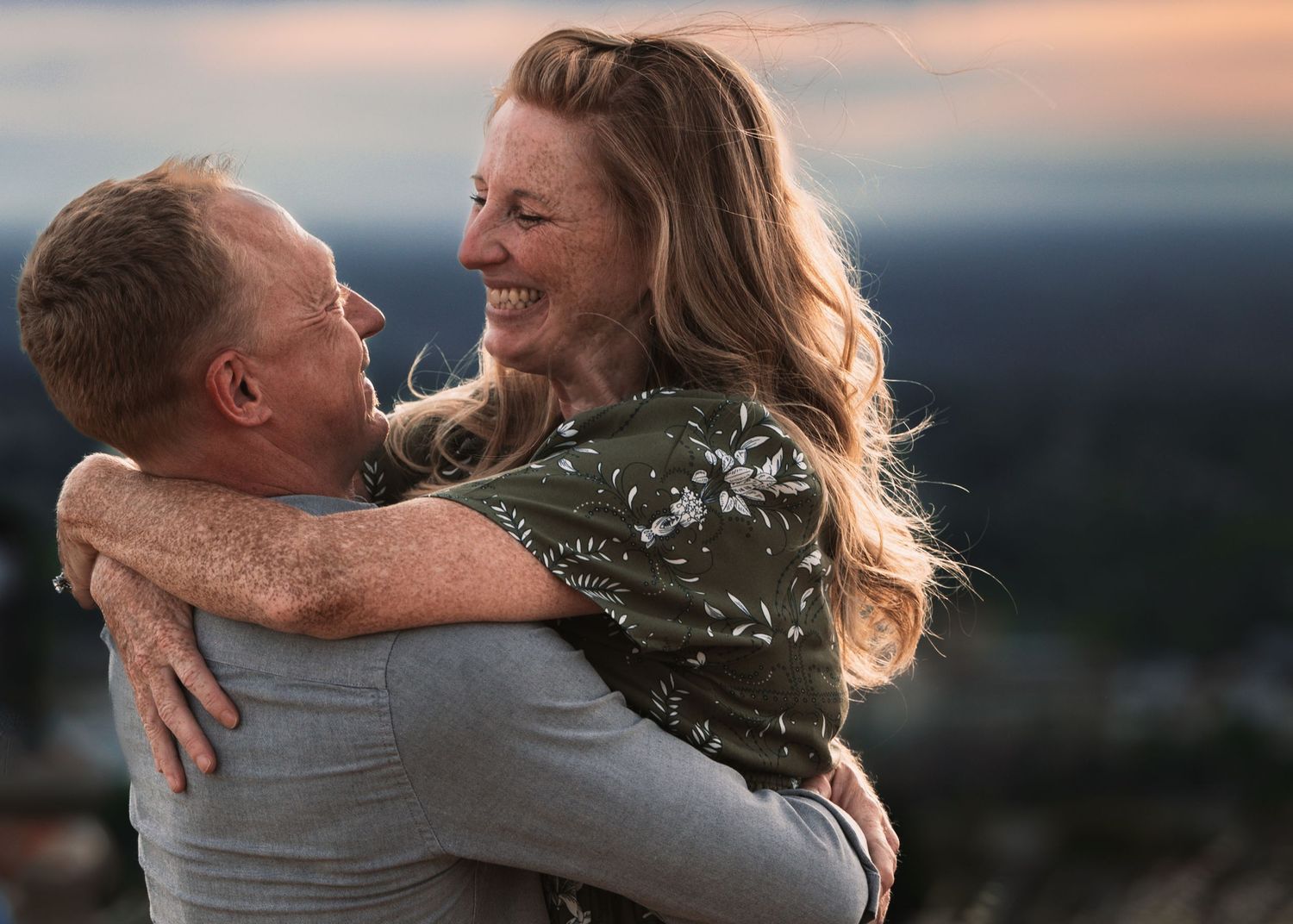 A joyful couple embraces outdoors at sunset, the smiling woman hugging a man against a scenic blurred background.