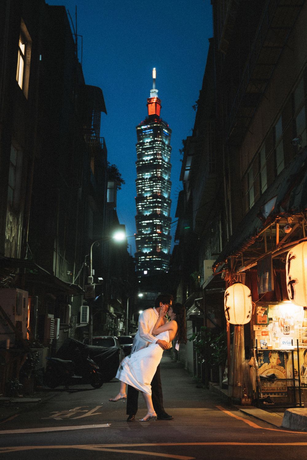 Couple embracing in a narrow alley at night with Taipei 101 tower glowing in the background.