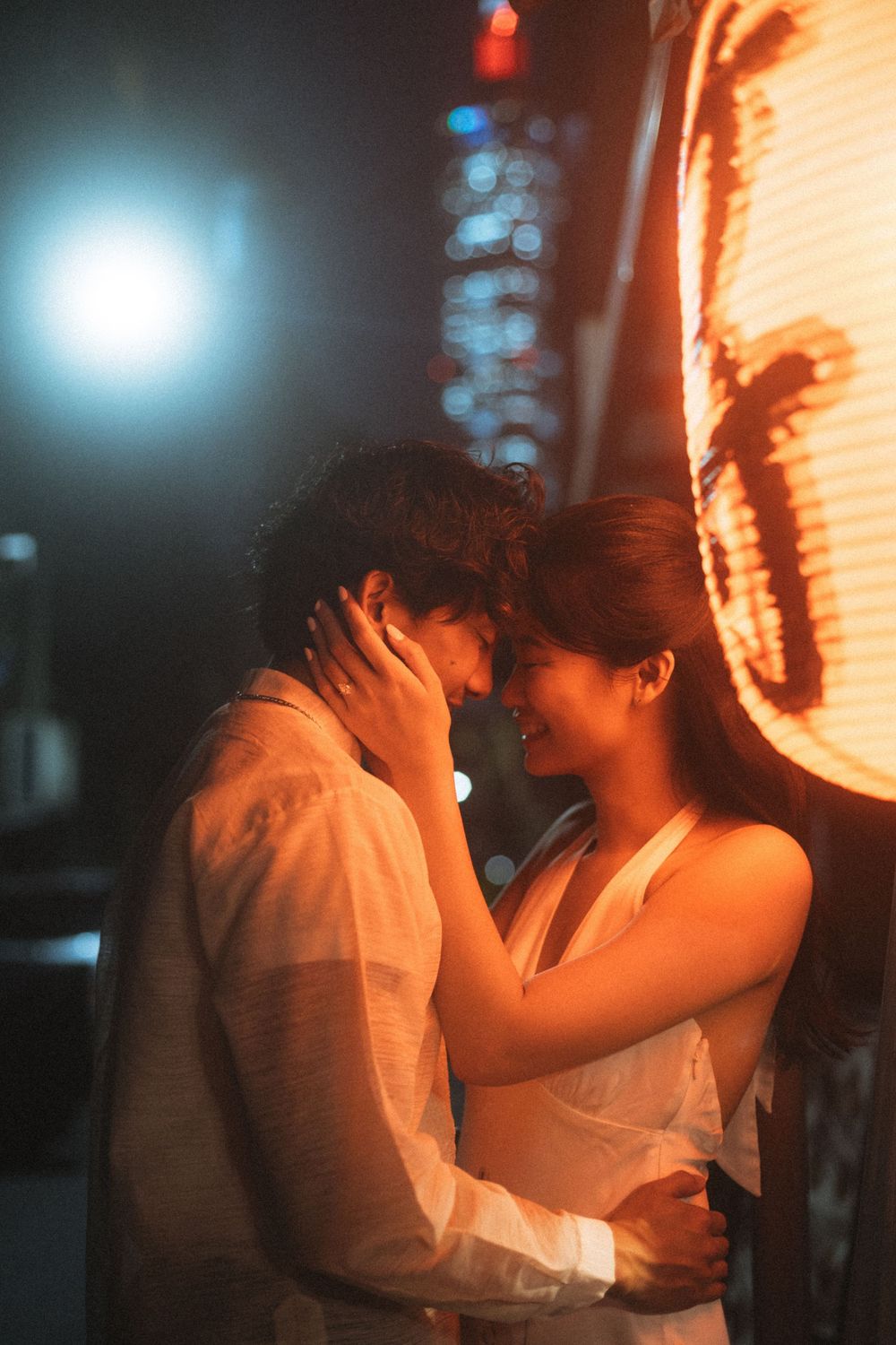 Romantic couple embracing near a glowing Japanese lantern at night with city lights in the background.