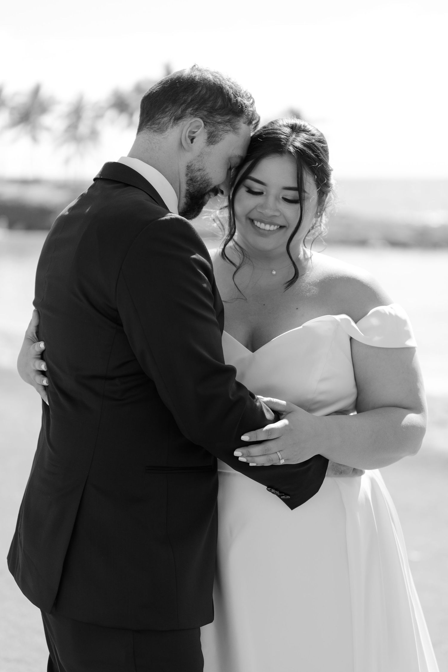 Black and white photo of a smiling bride in off-shoulder gown embracing a bearded groom in a suit on a beach.