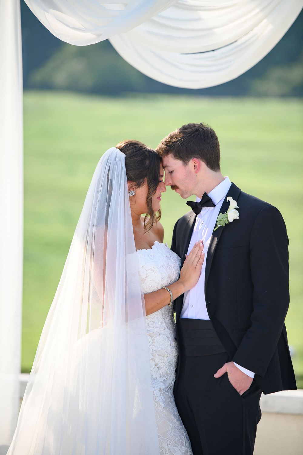Bride and groom share an intimate moment under white draped canopy, with lush green fields in the background.