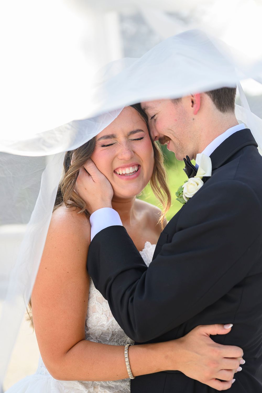 Bride and groom laughing joyfully under a wedding veil, sharing an intimate moment in formal attire.