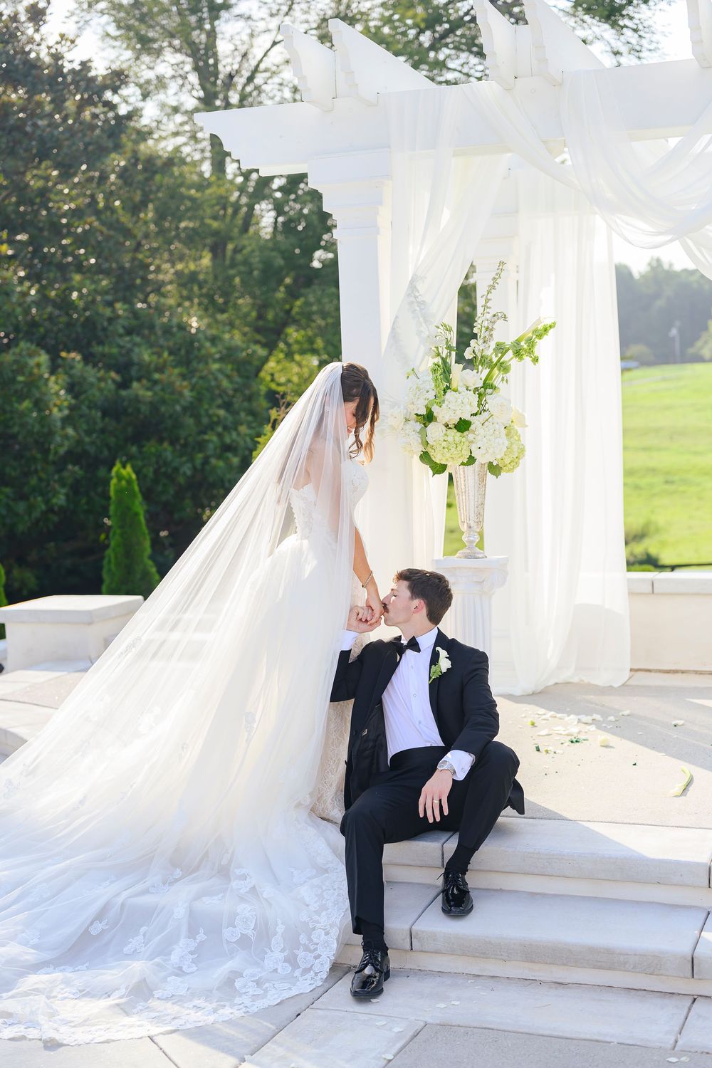 Bride and groom share a romantic moment on white steps near a floral arch outdoors.