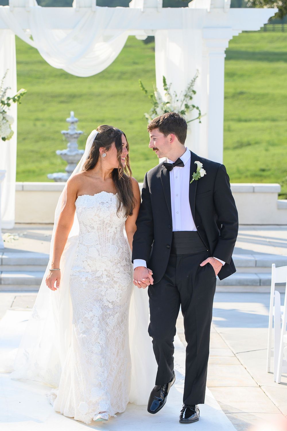 Bride and groom holding hands, smiling at each other outdoors near a white floral arch on their wedding day.