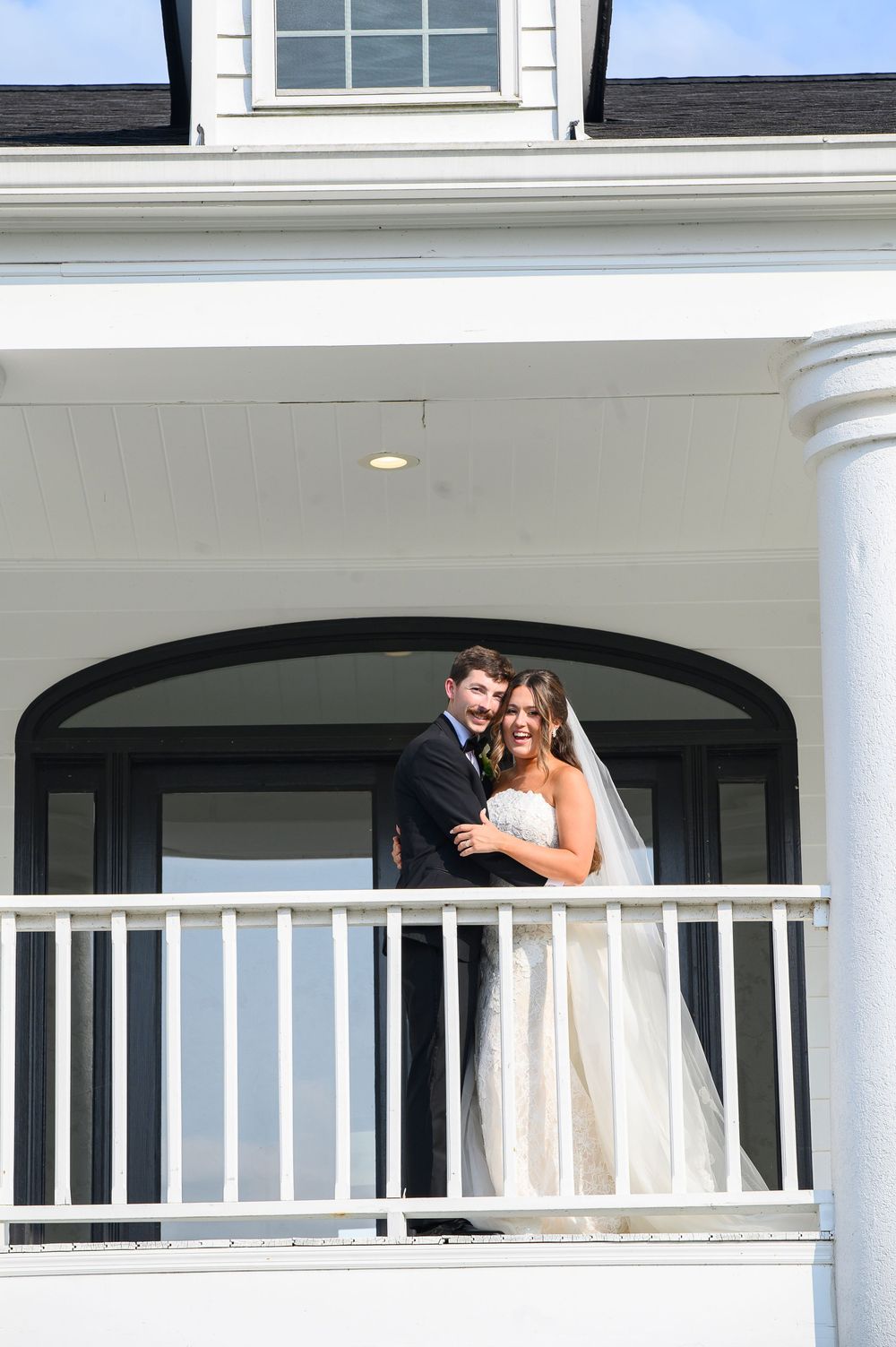 Bride and groom pose on white colonial porch balcony, bride in strapless gown with veil, groom in dark suit.