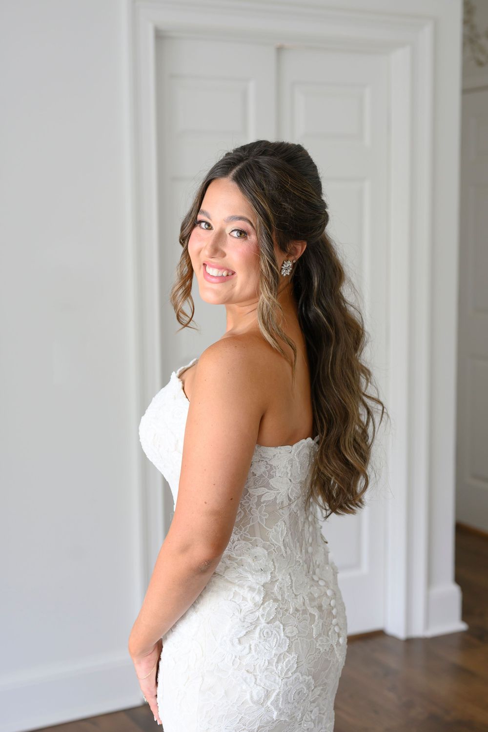 Bride in strapless lace wedding gown poses over shoulder, showcasing elegant half-up hairstyle and crystal earrings.