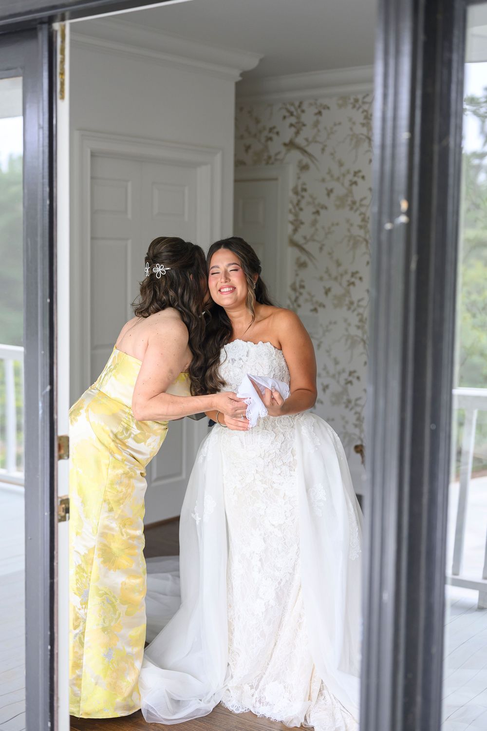 Bride in lace wedding gown smiles as bridesmaid in yellow floral dress helps her prepare in elegant bridal suite.