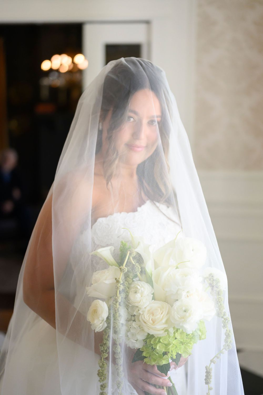 Radiant bride wearing a cathedral veil holds a lush white rose and green bouquet in a softly lit venue.