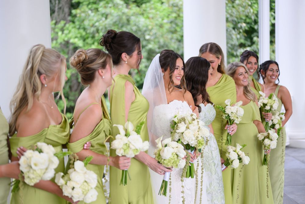 Bride in white gown surrounded by bridesmaids in sage green dresses holding white floral bouquets at elegant wedding.