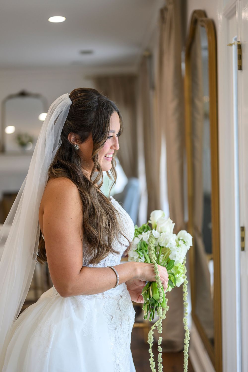 Smiling bride in white gown and veil holds elegant white floral bouquet with trailing greenery indoors.