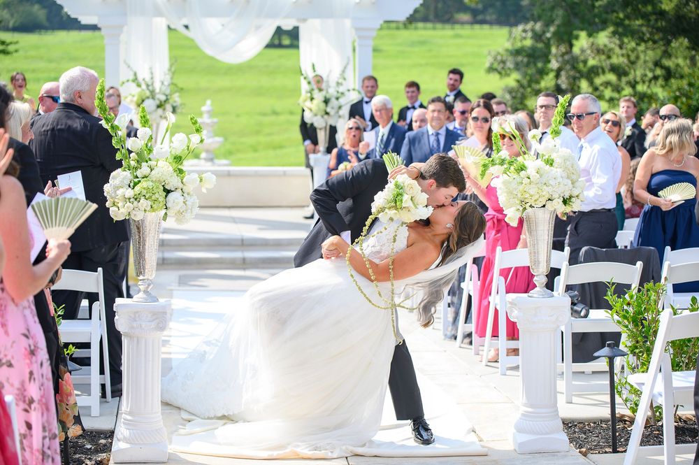 Groom dips bride for a kiss at outdoor wedding ceremony with white floral arrangements and guests seated on white chairs.