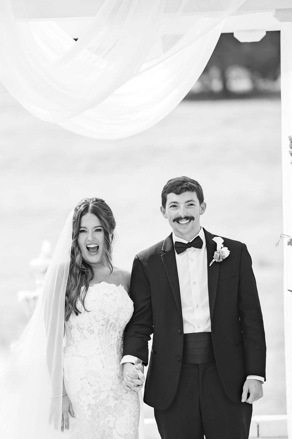 Black and white photo of a joyful bride and groom holding hands at their outdoor wedding ceremony.
