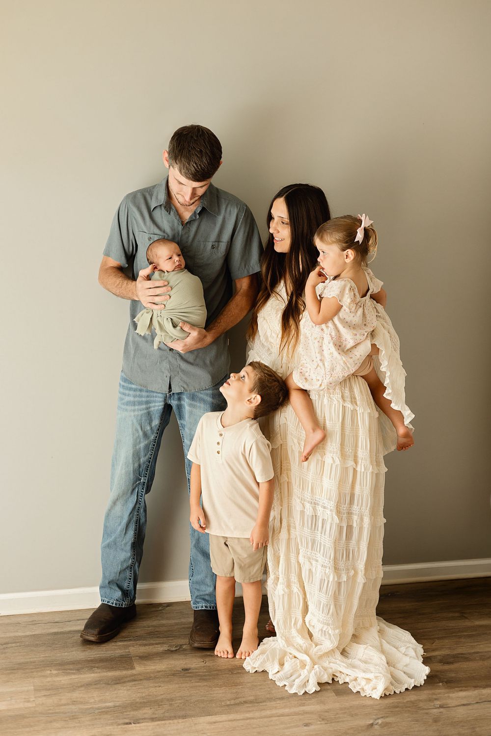 A young family of five poses indoors, with parents holding a newborn and toddler girl while a young boy stands between them.
