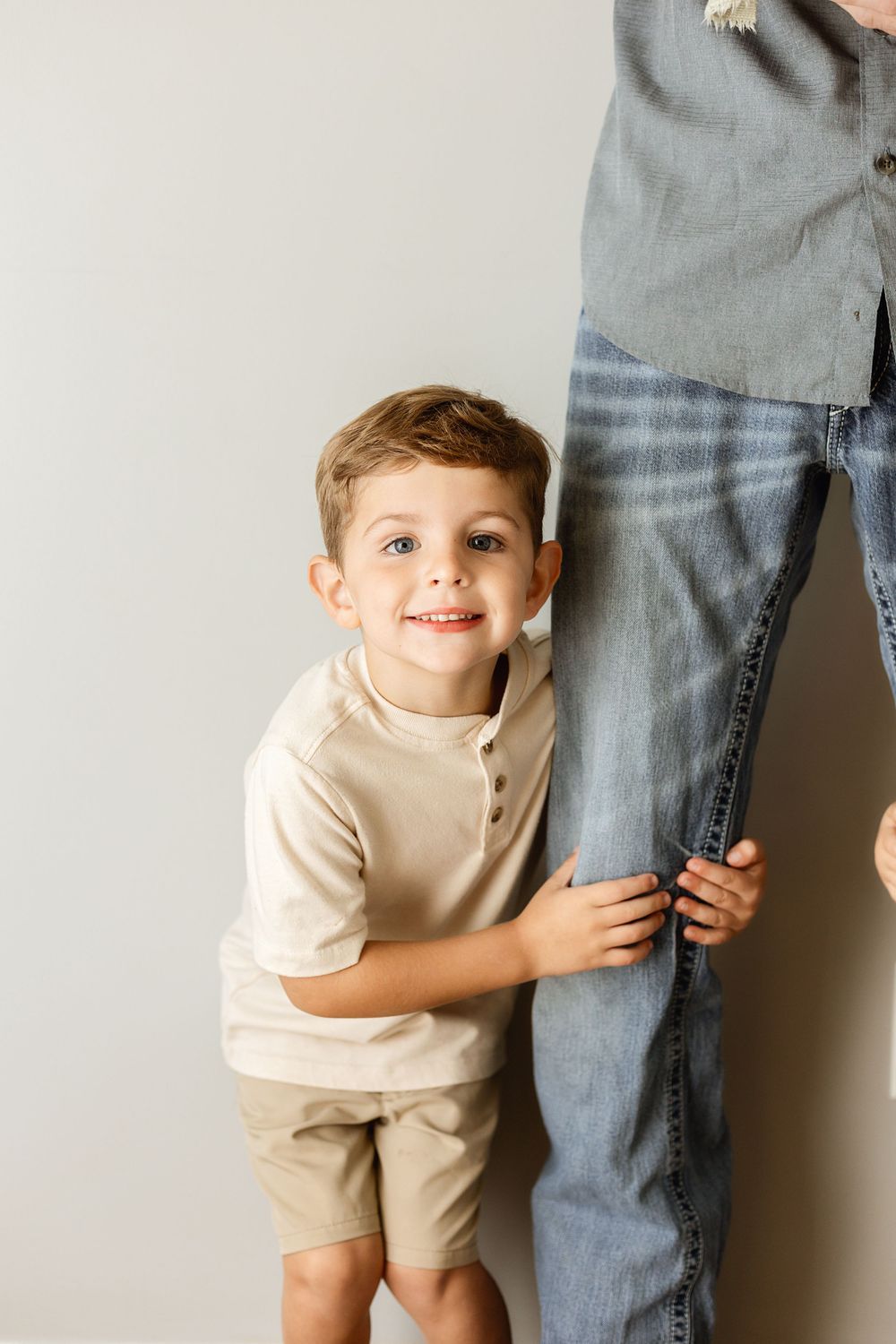 A smiling young boy in beige outfit hugs an adult's denim-clad leg against a light gray background.