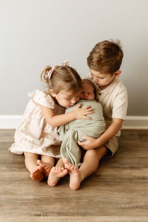 Two young siblings sit on a hardwood floor, lovingly cradling their swaddled newborn baby sibling.