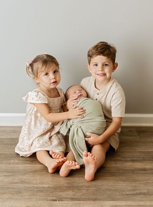 Two young siblings sit on hardwood floor holding a swaddled newborn baby, posing for a family portrait.