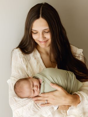 A smiling mother with long dark hair cradles her sleeping newborn wrapped in a sage green swaddle blanket.
