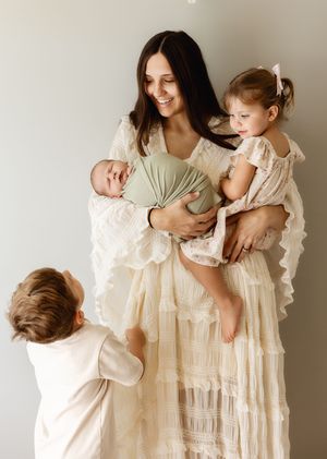 A smiling mother in a cream lace dress holds a newborn and toddler girl while a young boy reaches up to her.