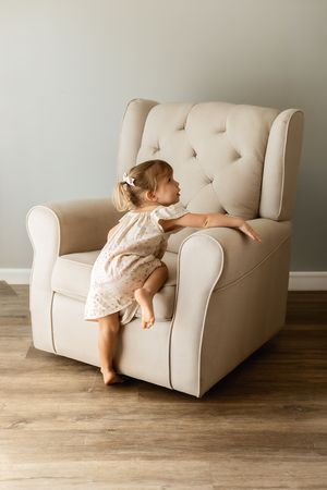 A toddler girl in a floral dress climbs onto a large beige tufted armchair on hardwood floors.