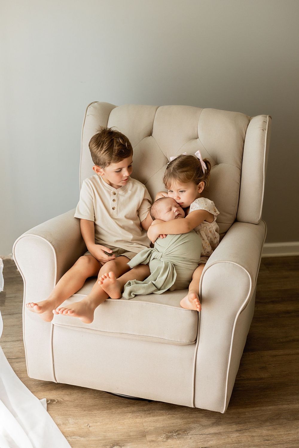 Two young children cuddle a newborn baby together in a cream tufted armchair, wearing neutral tones.
