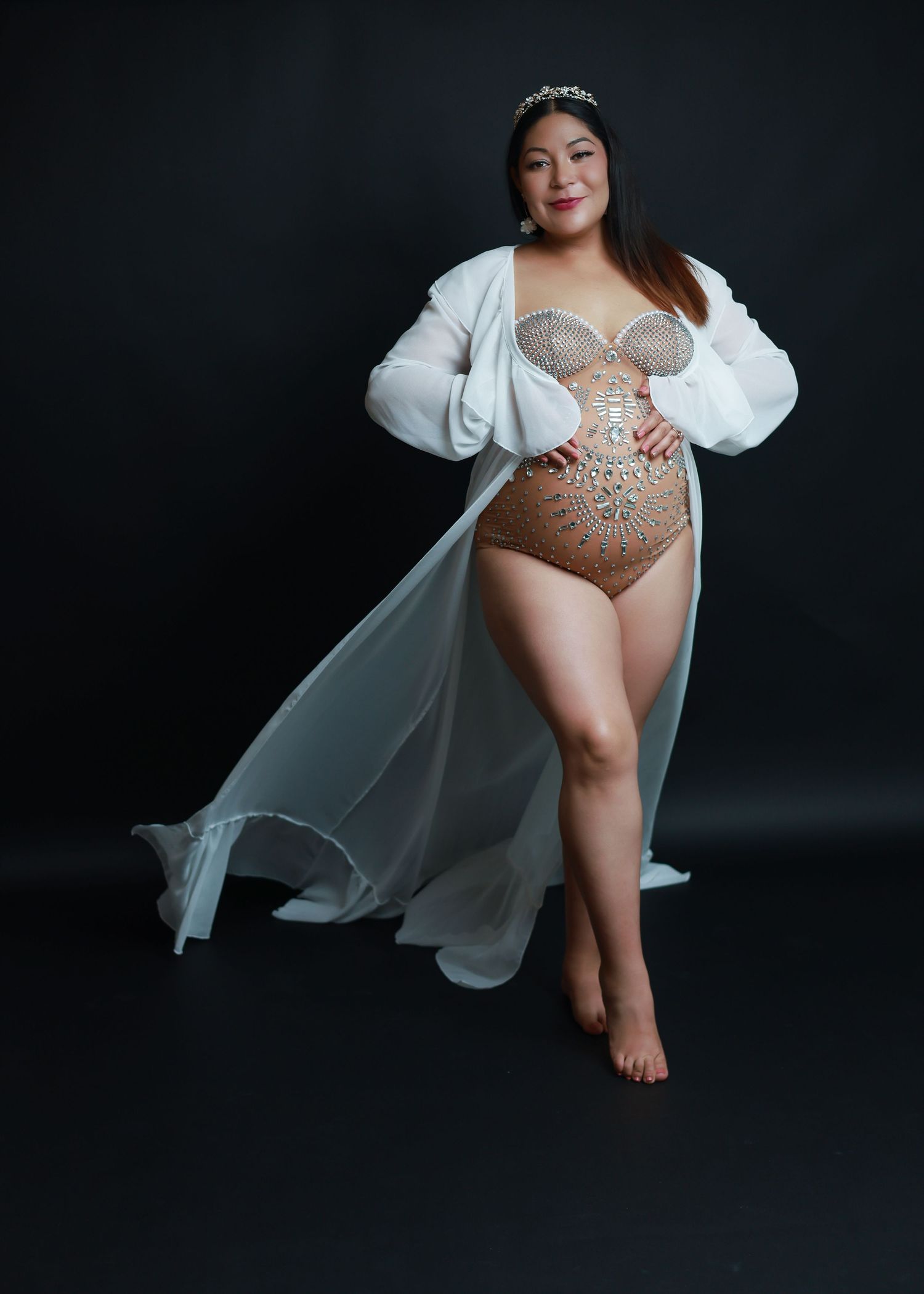 Pregnant woman in rhinestone bodysuit and white flowing robe with tiara poses gracefully against dark background.