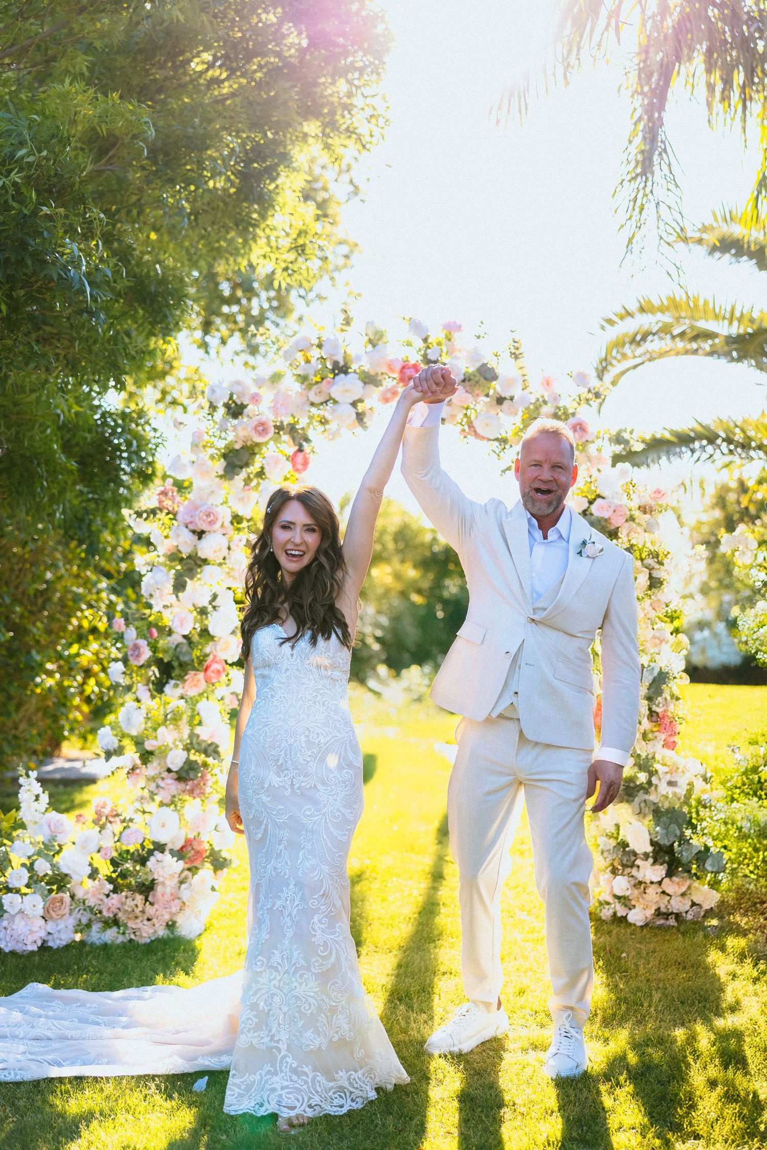 Bride and groom celebrating under floral arch at outdoor wedding ceremony on sunny day.