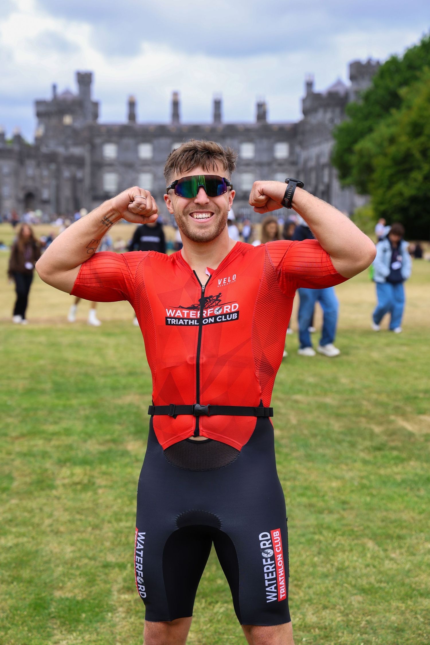 Athlete in Waterford Triathlon Club red wetsuit flexing muscles outdoors near historic castle grounds.