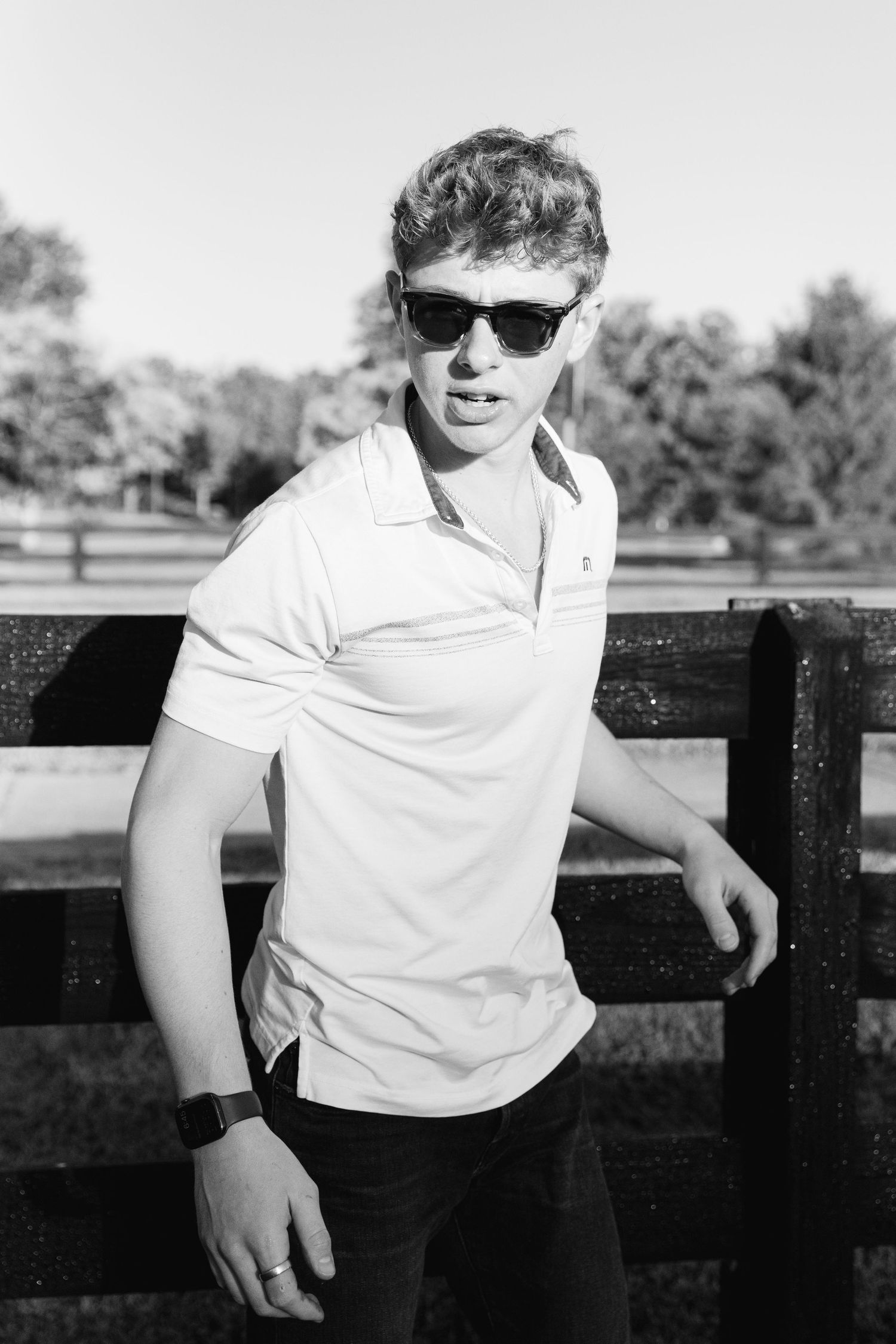 Young man in white polo shirt and sunglasses leans against wooden fence in black and white outdoor portrait.