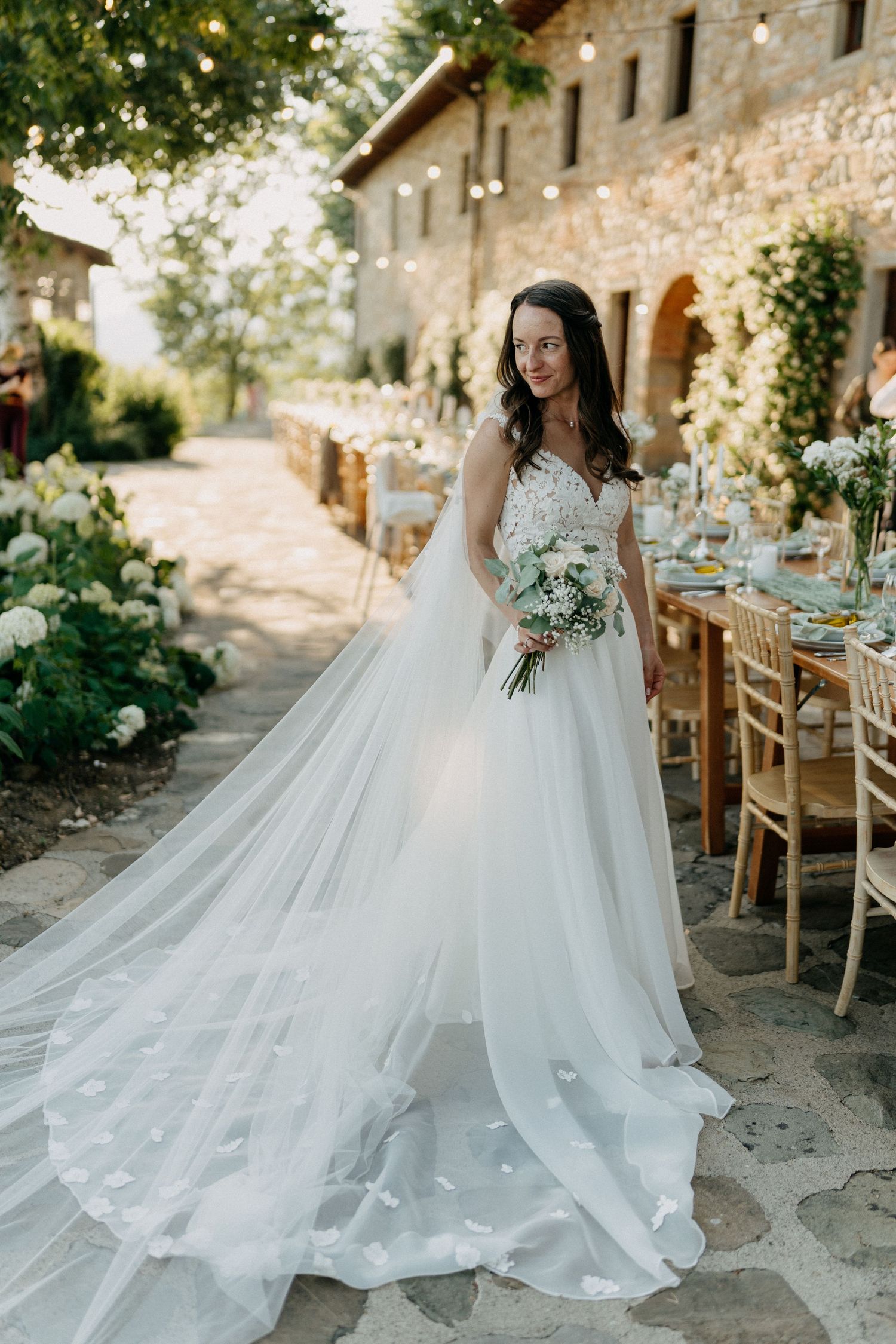 Bride in flowing white tulle gown holding bouquet at elegant outdoor Tuscan wedding reception venue.