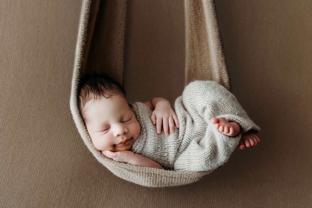 Newborn baby sleeping peacefully in a cozy knit hammock wrap, captured in a soft neutral-toned studio setting.