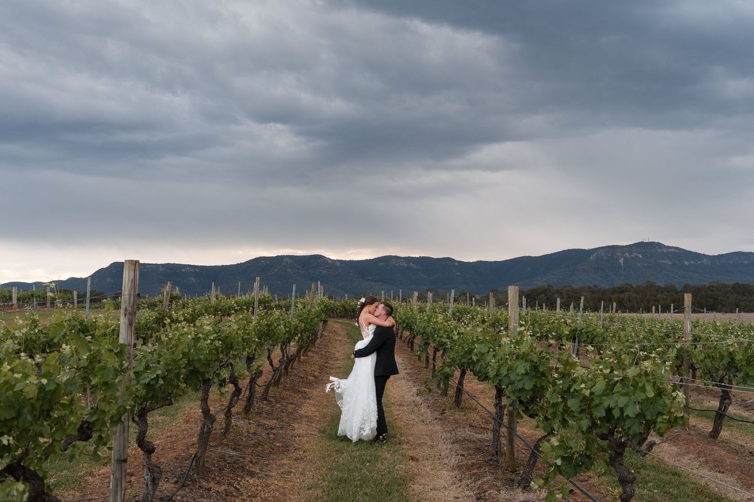 Bride and groom embracing in a vineyard with dramatic cloudy skies and mountain backdrop at sunset.