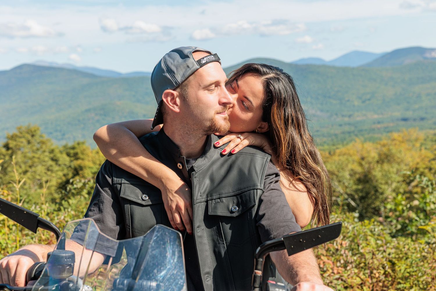 Woman kisses man on cheek while sitting on motorcycle with scenic mountain landscape behind them. North Conway NH