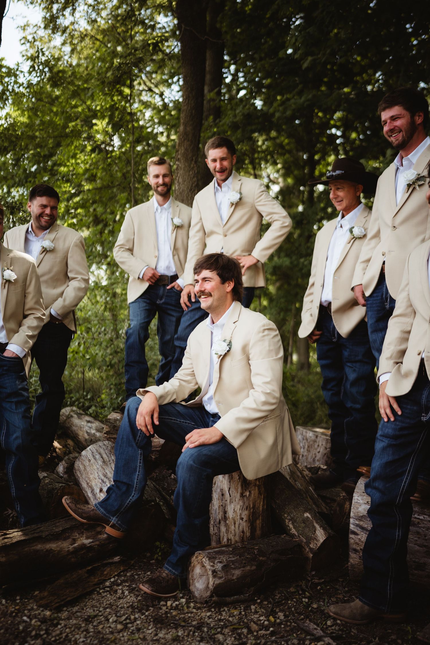 Groom and groomsmen in tan blazers and jeans pose outdoors on logs surrounded by trees at a rustic wedding.