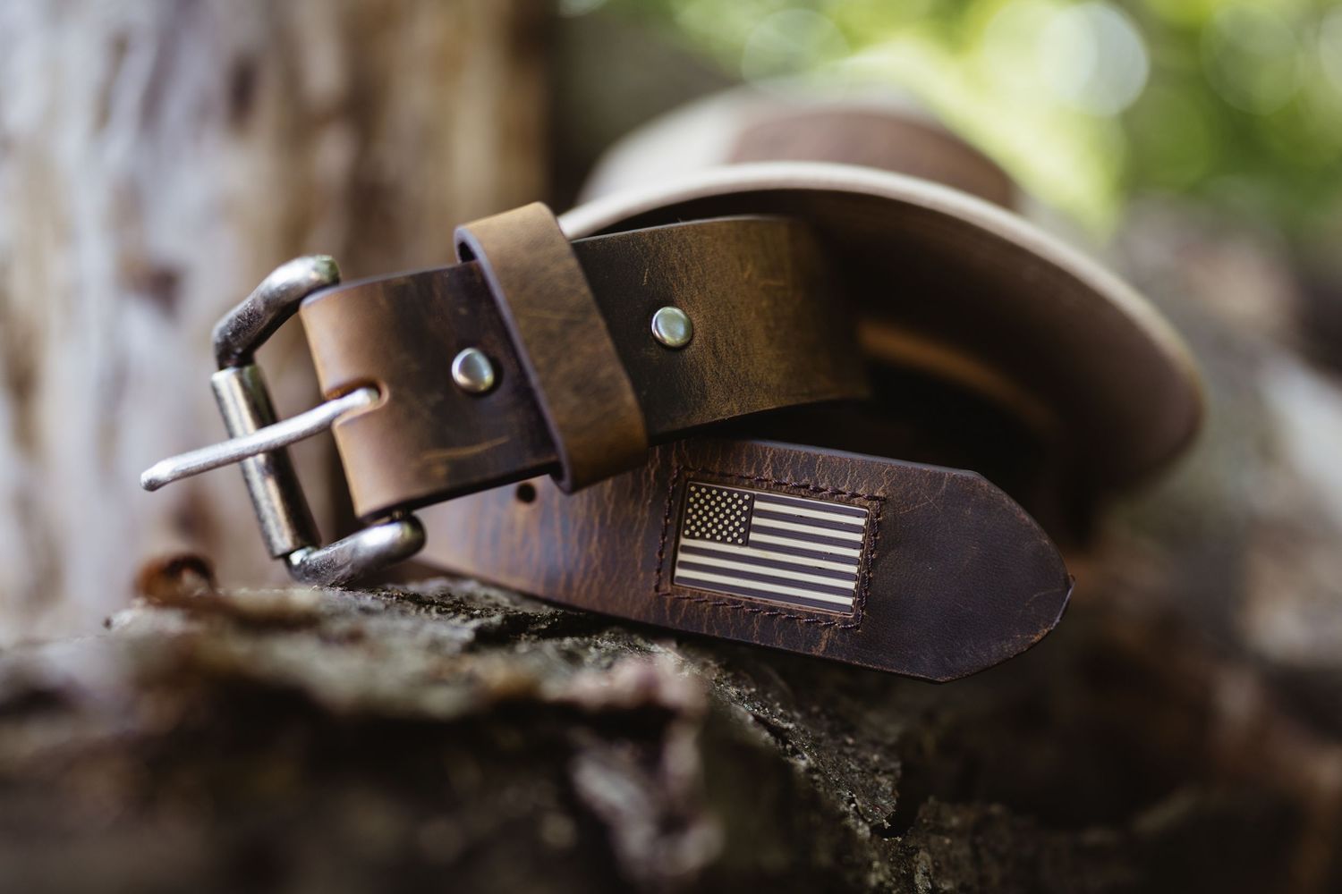 Brown leather belt with American flag embossed tip and silver buckle, displayed on rustic wood in natural setting.