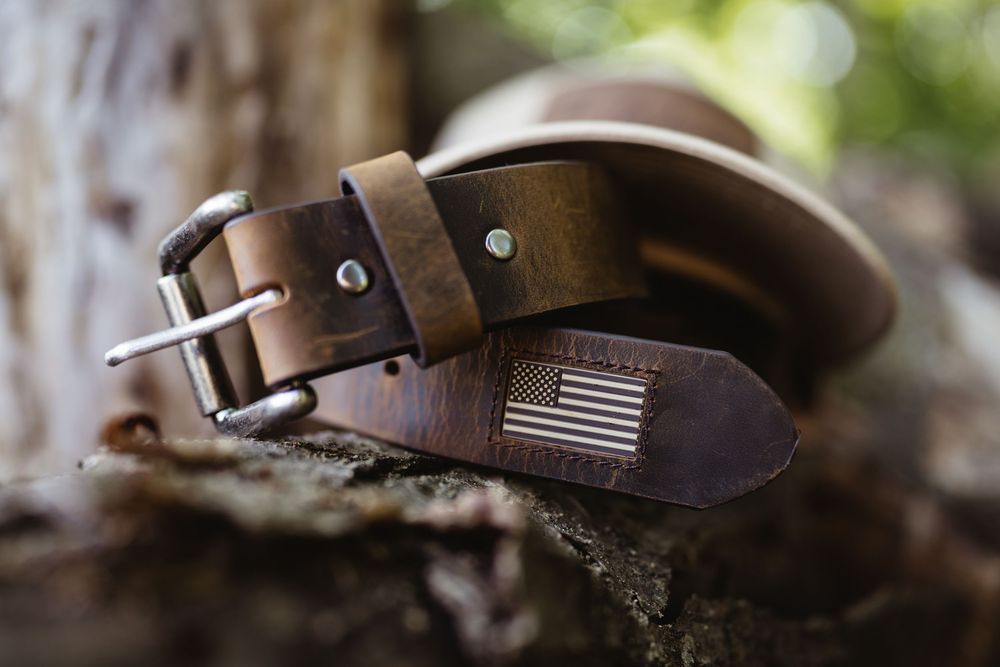 Brown leather belt with American flag embossed tip and silver buckle, displayed on rustic wood in natural setting.