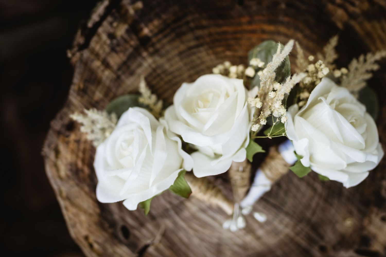 Three white roses with greenery arranged on a rustic wood stump, perfect for a wedding boutonniere display.