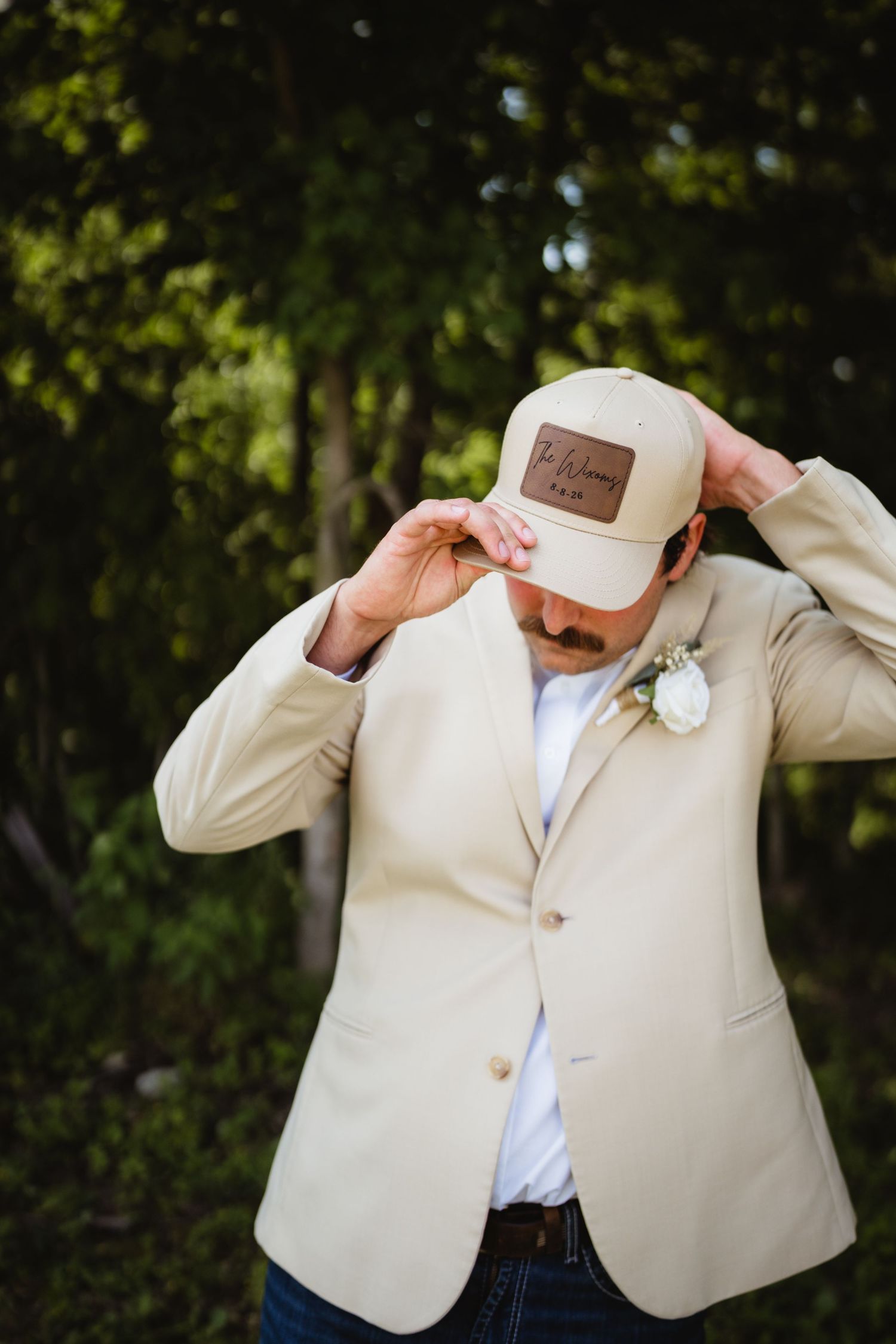 Groom in beige blazer adjusting personalized tan trucker hat, wearing boutonniere, outdoors with green trees.