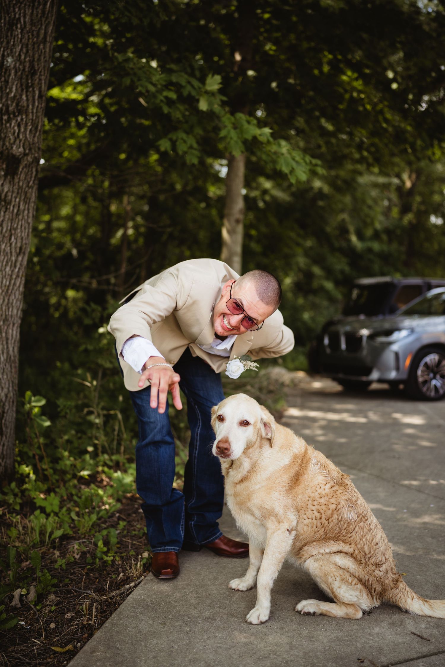 Smiling groom in tan blazer and boutonniere bends down to pet a golden Labrador on a tree-lined path.